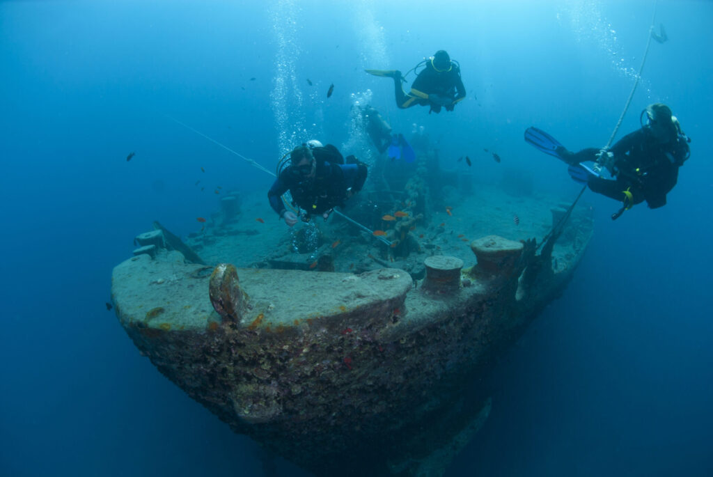 Silhouettes of scuba divers exploring the bow of a shipwreck.